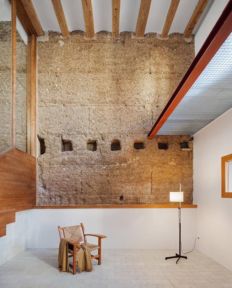 Patio interior con estructura de pared de madera original, revestimiento de gresite, acabados de madera, butaca de fieltro, lámpara de pie. La pared medianera de tapial coge un gran protagonismo en el nuevo lenguaje interior, puesto que técnicamente permite la regulación natural de la humedad del interior de la casa, y ofrece buenas prestaciones térmicas y acústicas.