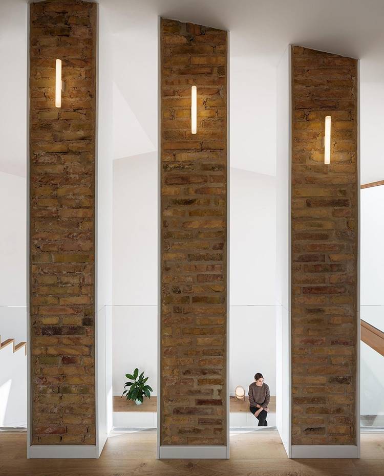 Pilares de obra vista con apliques,aperturas hacia nivel inferior, pilares recubiertos en blanco lado salón, . Las continuas aberturas verticales en la mitad superior de la pared que divide las dos plantas superiores favorece la conexión visual a la vez que dota dota de dinamismo al conjunto. 