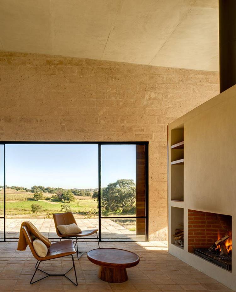 Revestimiento de gresite, módulo de obra con chimenea integrada, butacas de piel y mesilla frente a chimenea, apertura lateral hacia entorno natural . A nivel de suelo, dos grandes aperturas simétricas y acristaladas permiten que la casa se abra y se ventile de forma natural. Los cerramientos se protegen con paneles de madera correderos que encierran el espacio de forma compacta una vez no hay nadie habitándola. 