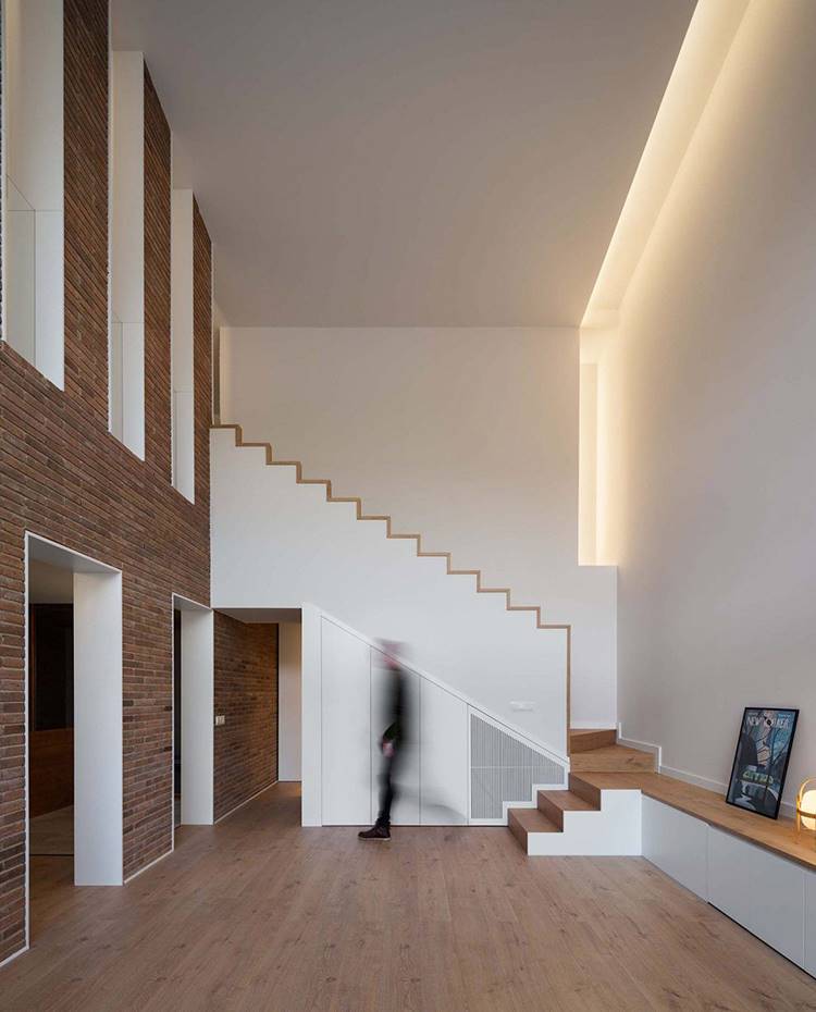 Salón a doble altura, escaleras de obra con revestimiento de madera, pared divisoria con aperturas revestida de ladrillo visto, mueble bajo frontal. En la concepción del proyecto se han combinado material original, texturas y colores con líneas limpias y depuradas para crear un nuevo lenguaje interior sorprendente. 