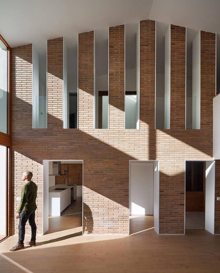 Zona de estar con muro separador entreabierto, muro revestido de ladrillo visto, pavimento de madera, cerramiento acristalado. La zona de día aglutina salón, comedor y cocina en un juego visual y espacial que termina abriéndose sobre la terraza exterior.