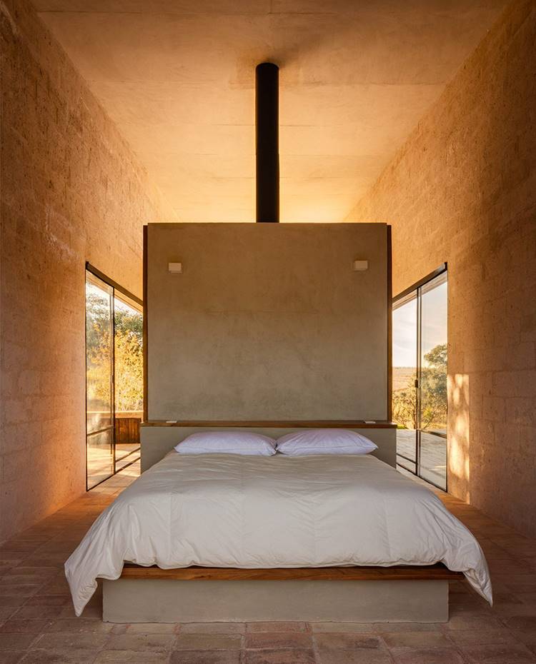 Zona dormitorio, cama central, módulo separador a modo de cabecero, apliques sobre estructura de obra. Tras la chimenea se ubica uno de los dos dormitorios de los que cuenta la casa. Todo ello en una disposición simple a la vez que efectiva, sin cerramientos interiores para favorecer la sensación de amplitud.