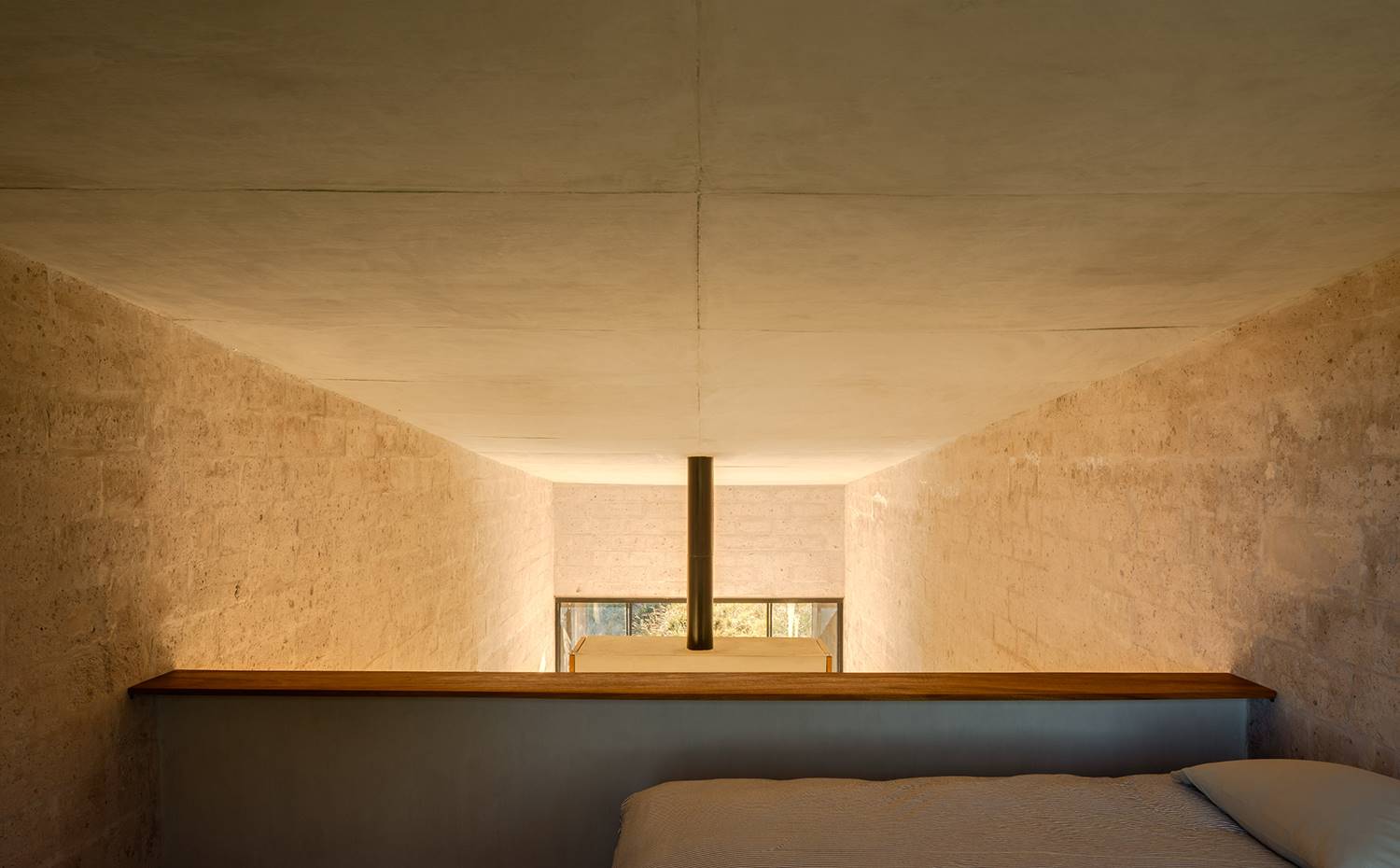 Dormitorio superior en altillo, con chimenea de fondo. Detrás de la chimenea, en el altillo adaptado en el extremo más elevado de la casa, se emplaza la segunda de las habitaciones, provista de una visión general del interior de la vivienda. Se accede a ella por una escalera de madera.