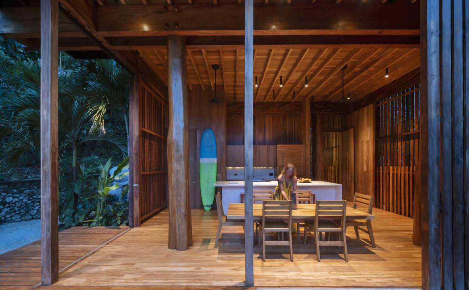 Cocina-comedor, todo revestido de madera, luces apliques techo, cocina en isla, tabla de surf, abierta al exterior.. La planta baja de la vivienda, a pie de bosque, es una zona diáfana con cocina y comedor, provista de una isla de trabajo que hace las veces de elemento separador. Todo ello revestido y con mobiliario de madera.