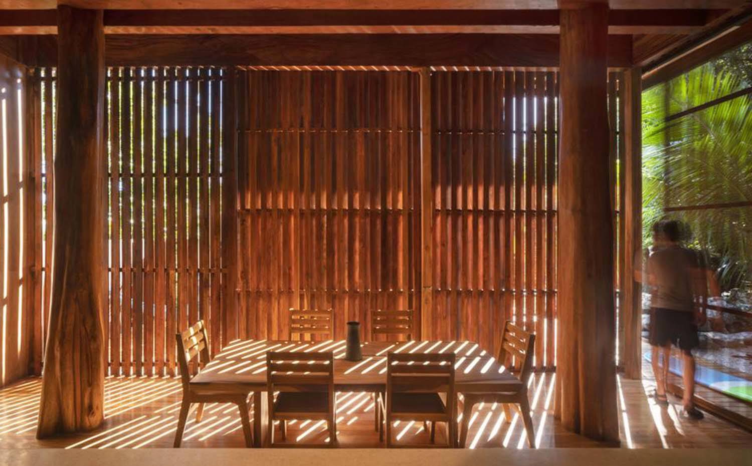 Comedor protegido del sol con puertas corrededar alistonadas, dos troncos a cada lado.. El comedor, totalmente de madera de teca, se abre y se cierra con puertas corrededar alistonadas que van de suelo a techo y ofrecen un acondicionamiento ambiental natural.