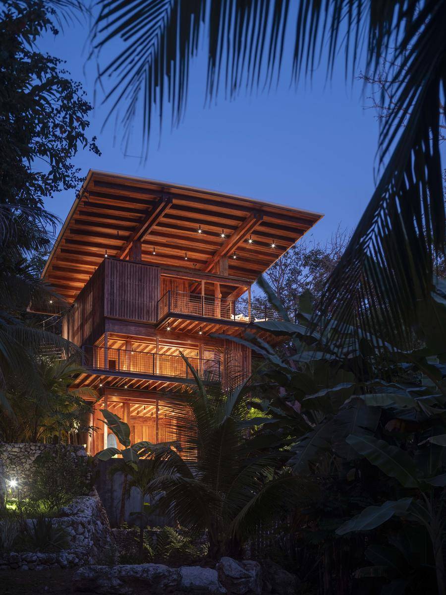 Exterior de la casa, vista nocturna de los tres niveles de la vivienda.. Diseñada como una cabaña para surfistas al aire libre, se organiza entorno a tres niveles: el primero, a ras de bosque; el segundo, en medio de la vegetación; y, el tercero, sobre las copas de los árboles y con vistas a la playa.  