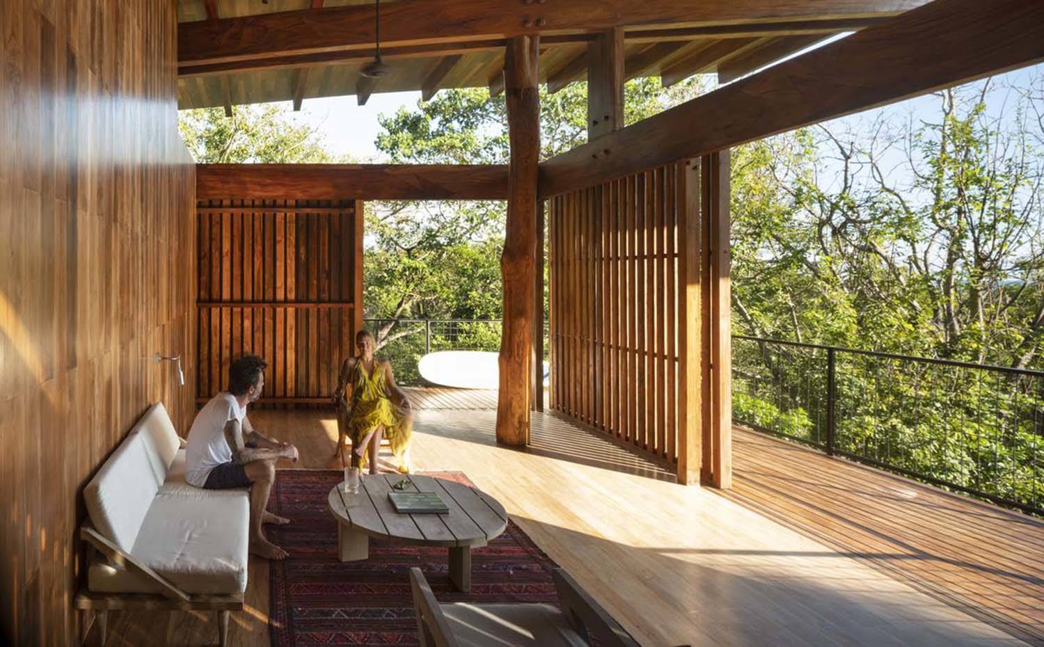 Zona de estar, nivel superior de la casa del árbol, sofá blanco, mesa de centro de madera, alfombra con motivos étnicos. estructura vivienda medera.. La segunda planta, destinada a zona de noche, cuenta con una sala de estar abierta, provista de poco mobiliario y bordeada por la terraza exterior. 