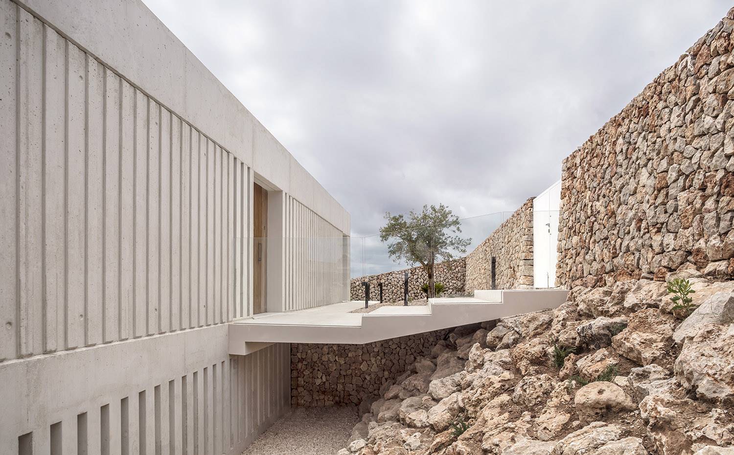 Entrada principal con pasarela de hormigón sobre foso, muro de piedra, . Para combatir la pendiente del lugar, la vivienda se posiciona en el extremo superior del terreno y se habitúa el acceso principal por la planta superior en un juego armónico entre hormigón, madera, cristal y piedra. 