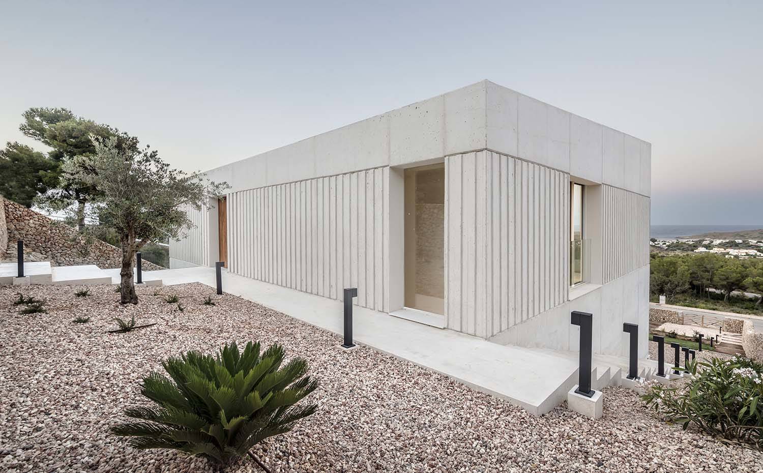 Fachada de hormigón alistonada, aperturas de cristal, escaleras de accesos a los diferentes niveles, zona ajardinada con piedras, árboles y arbustos.. El hormigón moldeado in situ fue elegido para la fachada puesto que es un material que no precisa mantenimiento y envejece de forma óptima teniendo en cuenta el clima de Menorca, con altos índices de humedad y fuertes niveles de sal. 