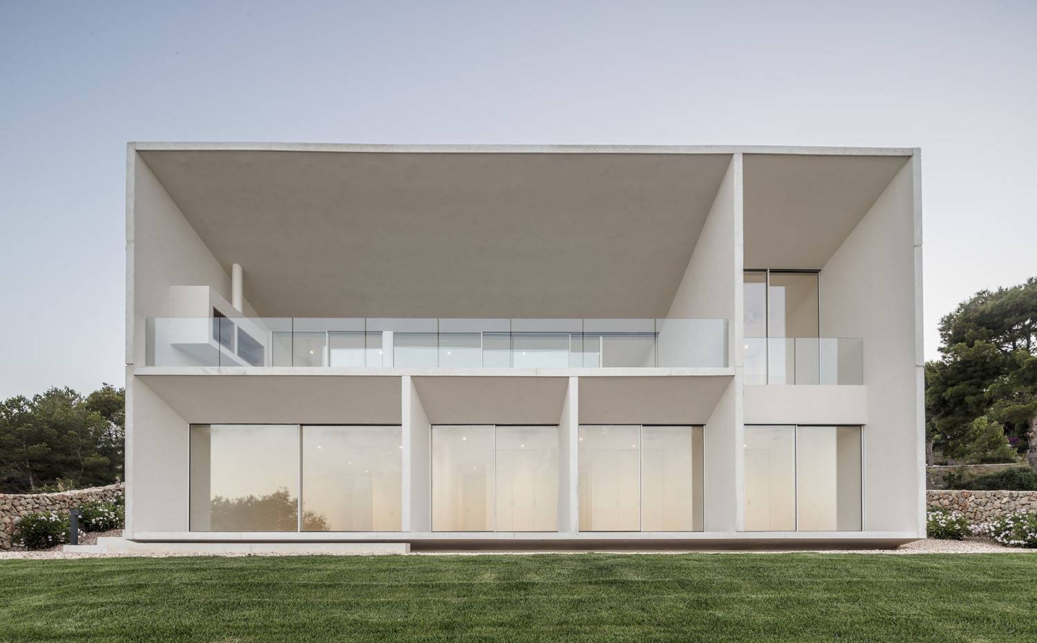 Frontal exterior, jardín con césped, cuerpo restangular con particiones muy lineales.. Grandes aberturas a lo largo de la fachada de hormigón enmarcan amplias vistas hacia el Mediterráneo en esta vivienda en la costa de Menorca.