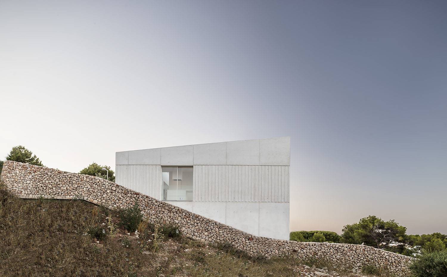 Vista exterior de la vivienda con apertura hacia la cocina, techo inclinado, rodeada con murete de piedras. La vivienda, proyectada por el estudio de arquitectura Nomo Studio, con sede en Barcelona, ocupa una empinada parcela en Coves Noves, en la costa noreste de la isla.