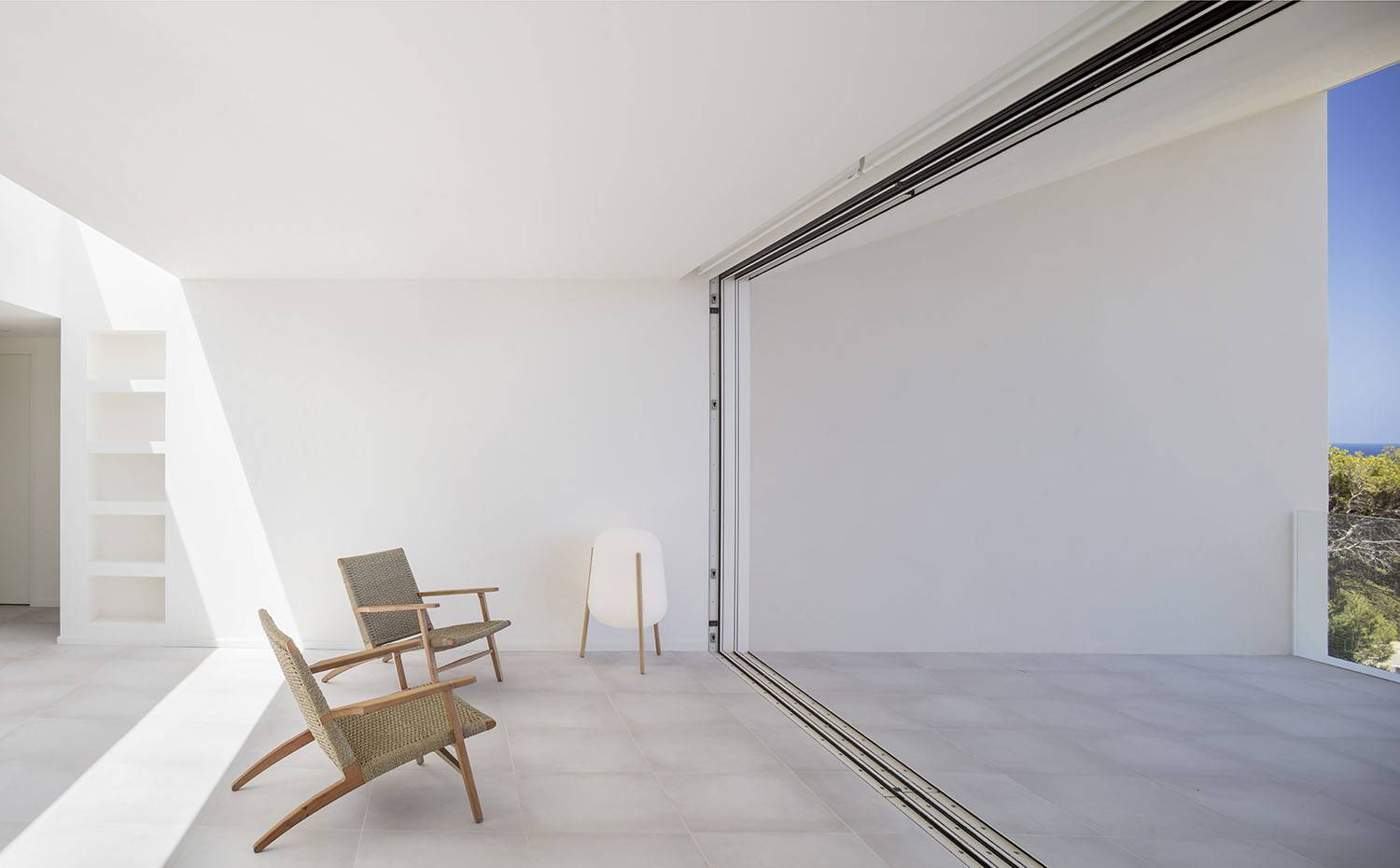 Zona de estar interior con suelo de gresite, cerramiento acristalado, butacas de fibra y madera, lámpara de pie con pies de madera. El empleo de los mismos materiales para las zonas interiores y exteriores de la vivienda hace que ambas se fusionen física y visualmente adaptándose a las necesidades de cada momento, 