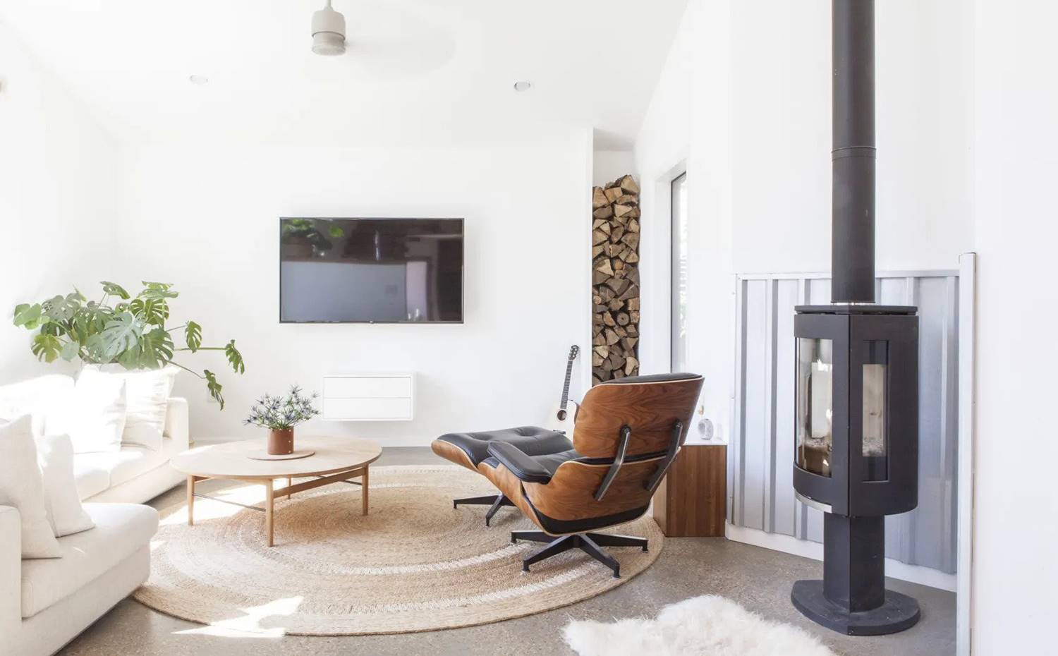 Bucata con reposapies de madera y piel, chimenea de hierro, columna de troncos de madera, ventilador en techo, sofá blanco, mesilla de centro de madera. El salón mezcla elementos de inspiración industrial con otros de estética más cálida y acogedora. El sillón Eames Long atrae las miradas en un espacio que evoca paz y sosiego.