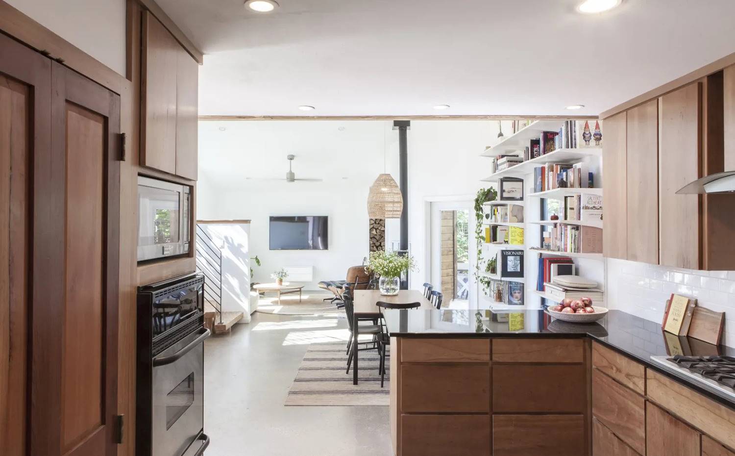 Cocina abierta a comedor y salón, con módulo separador en península, mobiliario en madera con tiradores intergrados. El mobiliario de cocina, en madera oscura y con tiradores intergrados, presenta una disposición en foma de L que asoma sobre el comedor y el salón, acompañada de otro frente de almacenamiento donde se integran los electrodomésticos..