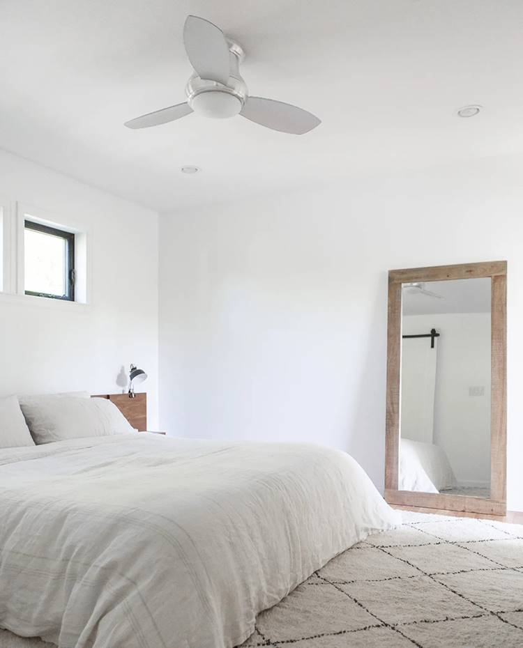 Dormitorio con moqueta blanca a rayas negras, textil de hogar blanco, espejo apoyado a suelo con marco de madera, ventilador en techo. El dormitorio principal, totalmente austero y puro, proyecta una gran sensación de serenidad. La fuerte entrada de luz a ambos lados de la estancia, junto a la gran alfombra mullida y la dominante blanquecina, hacen de la estancia un paradigma de paz y reposo.  