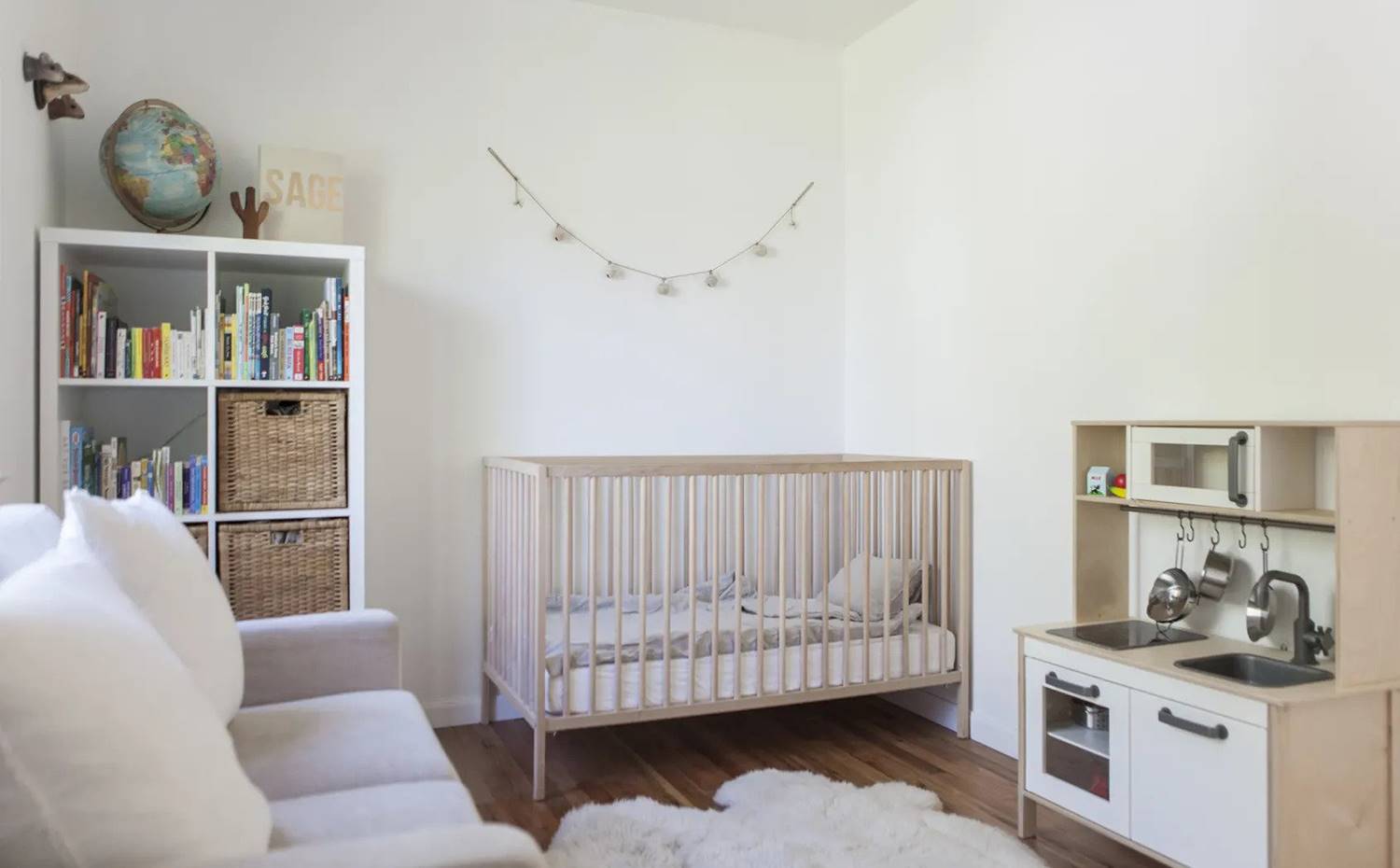 Dormitorio infantil con cuna de madera, librería blanca, cocina de juguete de madera, sofá y alfombra de pelo largo. El dormitorio infantil está equipado con elementos funcionales que pueden adaptarse a todo tipo de ambientes, con la finalidad de ir modificando la estancia a medida que la pequeña vaya creciendo.