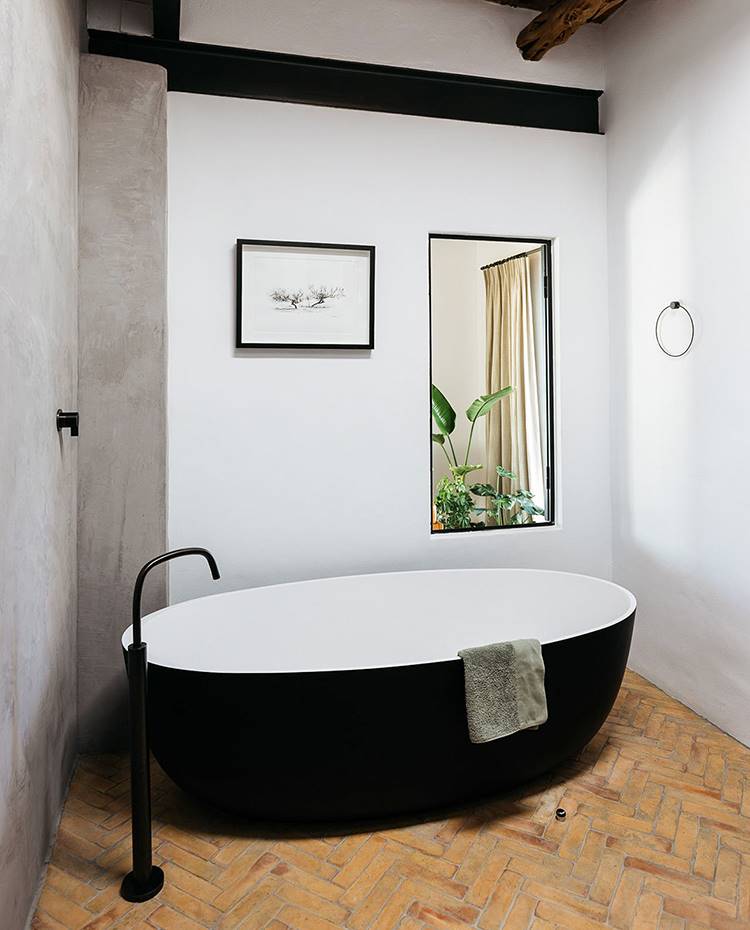 Baño con suelo cerámico en espiga, bañera exenta negra, grifería de pie negra, apertura a dormitorio. El cuarto de baño dispone de bañera independiente y ventanas con vistas a la naturaleza. El suelo en espiga hecho de baldosas de terracota es una reinterpretación moderna del diseño tradicional.
