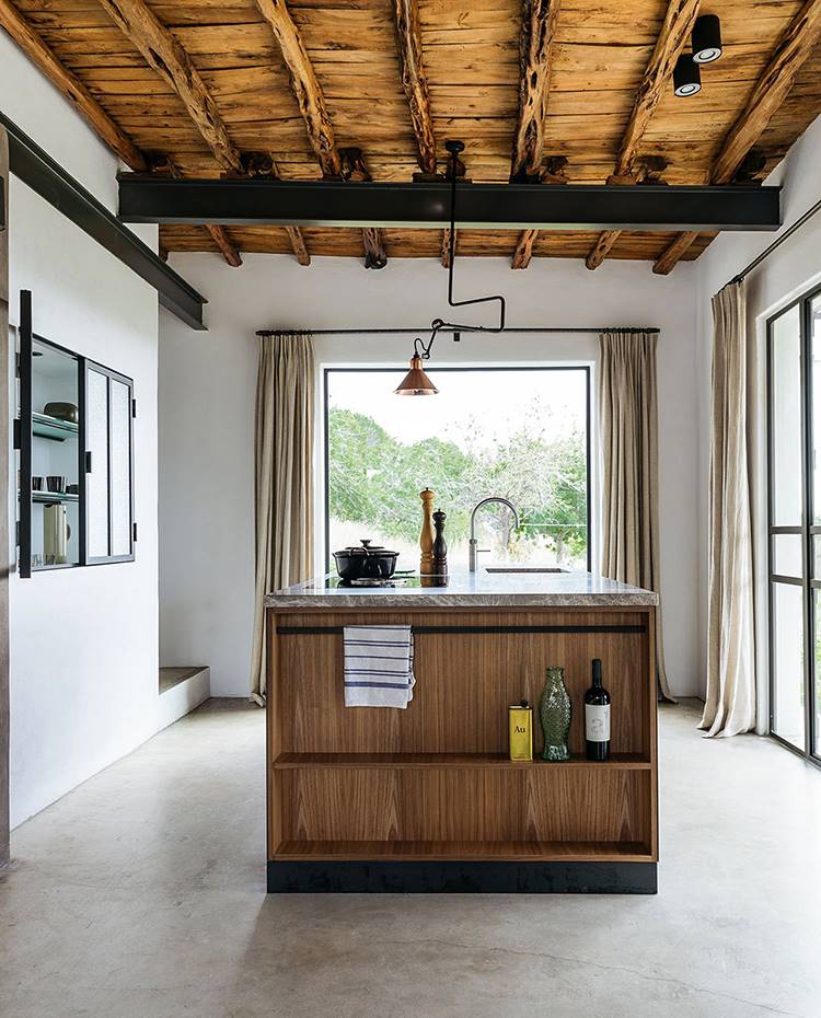 Cocina en isla, suelo de hormigón, vigas de hierro, techo de madera, vitrina en obra con cerramiento de cristal, luz suspendida con flexo.. La cocina de acero, hecha a medida, presenta una encimera de mármol y un armario con marco de acero y cerramiento acristalado que sigue el mismo patrón que las ventanas de la casa. 