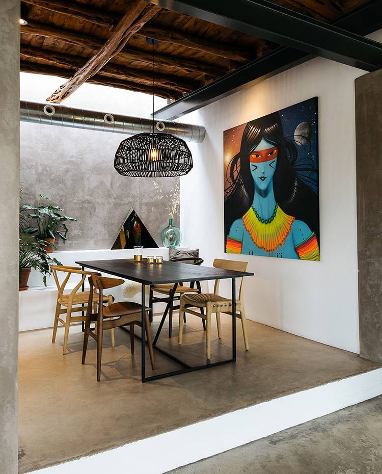 Comedor elevvado con suelo de hormigón, lámpara suspendida con pantalla de fibra, mesa con estructura metálica y sobre de madra, sillas de madera, conducto ventilación a vista. El comedor elevado, junto al salón, se acompaña de un banco de madera y una atractiva luz natural procedente de la ventana de vidrio fija en el techo, generando una agradable atmósfera.