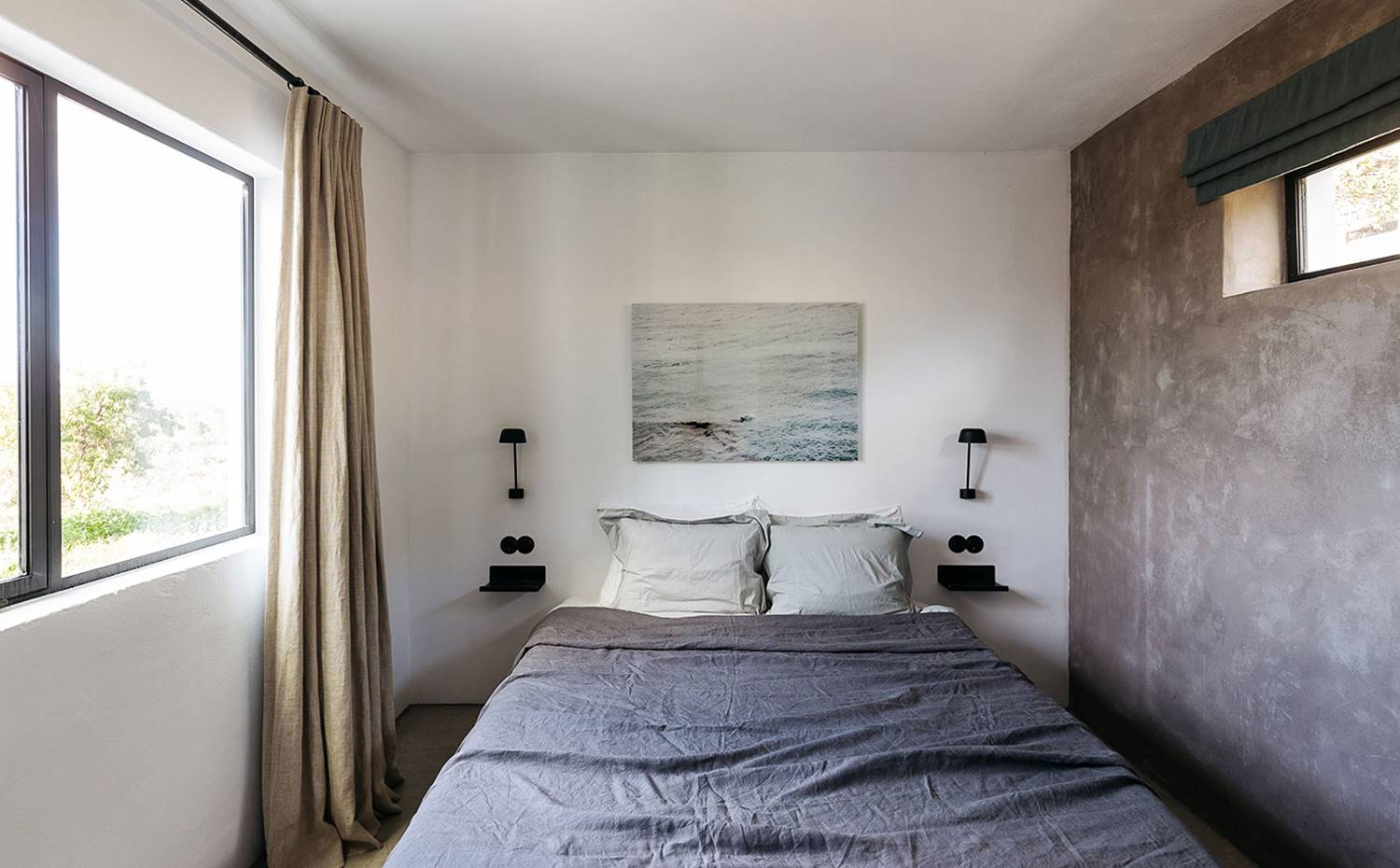 Dormitorio con lencería de hogar de lino, pared en gris, apliques negros, baldas a ambos lados de la cama, aperturas a ambos lados. La vivienda proyectada por Jurjen van Hulzen se extiende hacia el exterior con esta terraza que asoma al valle y regala vistas panorámicas de la vegetación propia de la zona.
