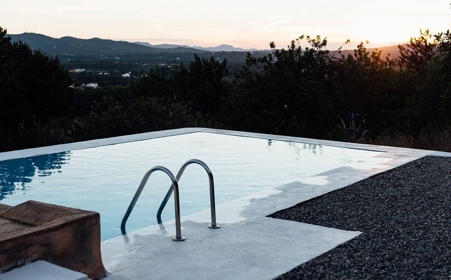 Piscina desvordante sobre paisaje natural, anochecer. La piscina exterior está rodeada por una hermosa terraza de guijarros que respeta el entorno natural del valle en el que se ubica la vivienda, al norte de la isla de Ibiza. 