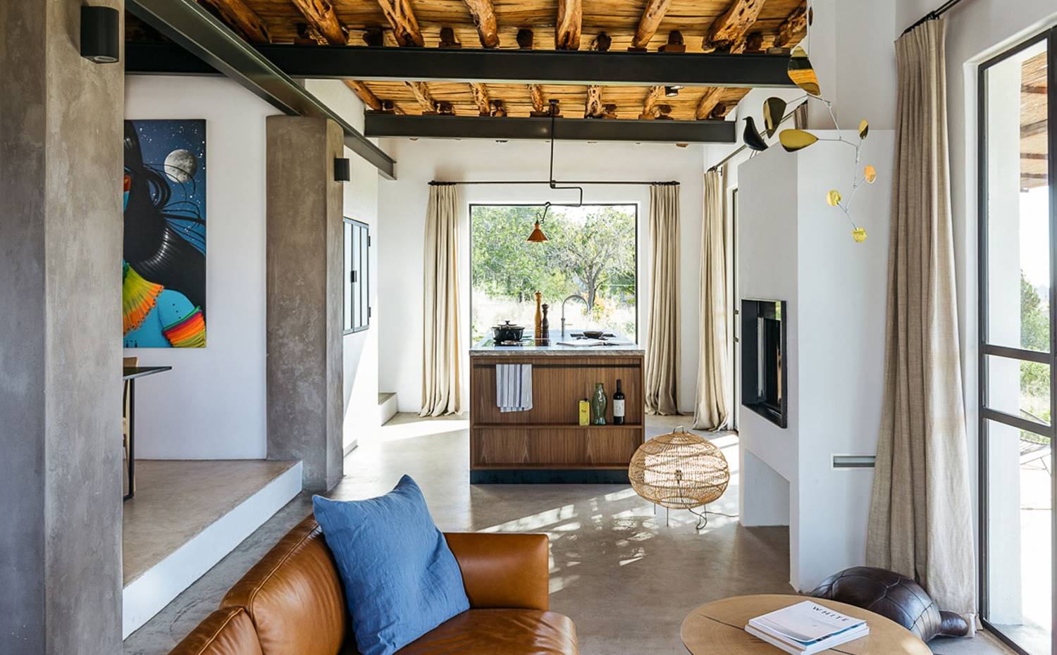 Zona comedor y salón, lámpara de suelo de fibra, sofá de piel marrón, cojín azul, revestimientos y suelo de hormigón, cortinas de lino en cerramientos, vigas de hierro. Los suelos de hormigón y las paredes de piedra con yeso y barro realzan la belleza natural del techo de madera a base de vigas Sabina. Todos los materiales utilizados en el proyecto son locales, excepto el acero con recubrimiento en polvo utilizado en los marcos de ventanas, armarios y puertas.