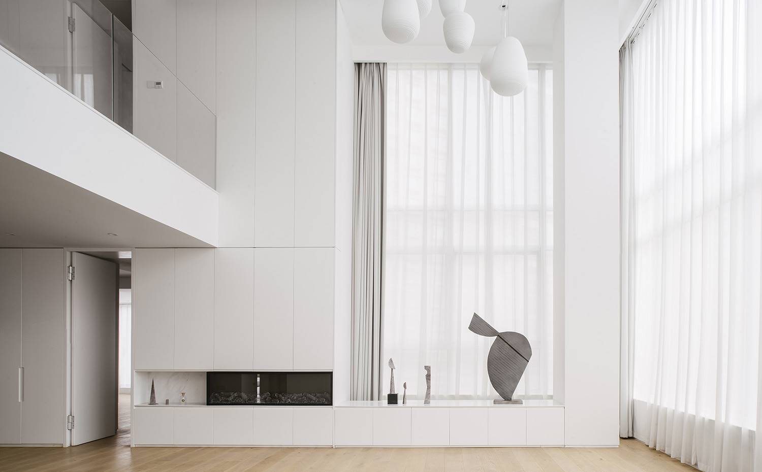 Sala de estar de doble altura, mobiliario a medida blanco con chimenea integrada, suelo de parquet, barandilla de cristal, lámparas suspendidas bvaladas. Con una propuesta de diseño simple y puras, Office ZHU ha logrado ofrecer un espacio cálido y acogedor para toda la familia, donde se impone el color blanco y las líneas depuradas. Todo ello acompañado de pequeñas esculturas y pinturas como toque personal final por parte del cliente.