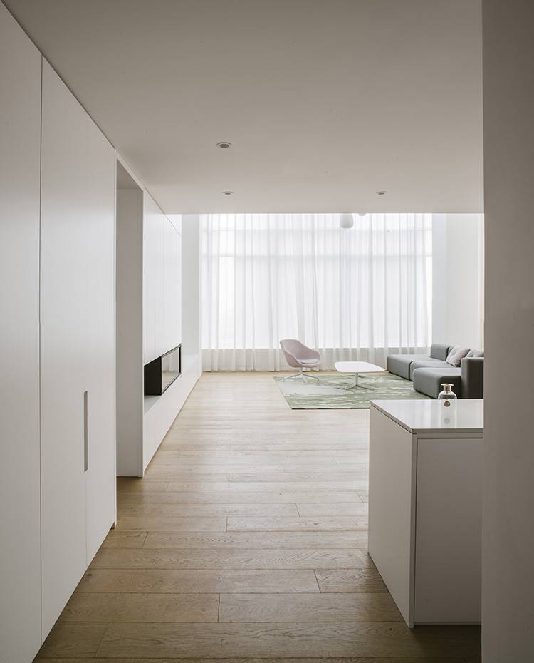 Vista desde entrada, isla de zona de vinoteca, armarios almacenaje en blanco, salón con amplio sofá y butaca en rosa, mesa de centro con sobre de madera, cortinas frente a ventanales.. Tras cruzar la puerta de entrada, una atmósfera espaciosa y muy luminosa da la bienvenida y sirve de base para todo el proyecto de reforma llevado a cabo por el estudio de arquitectura Office ZHU.