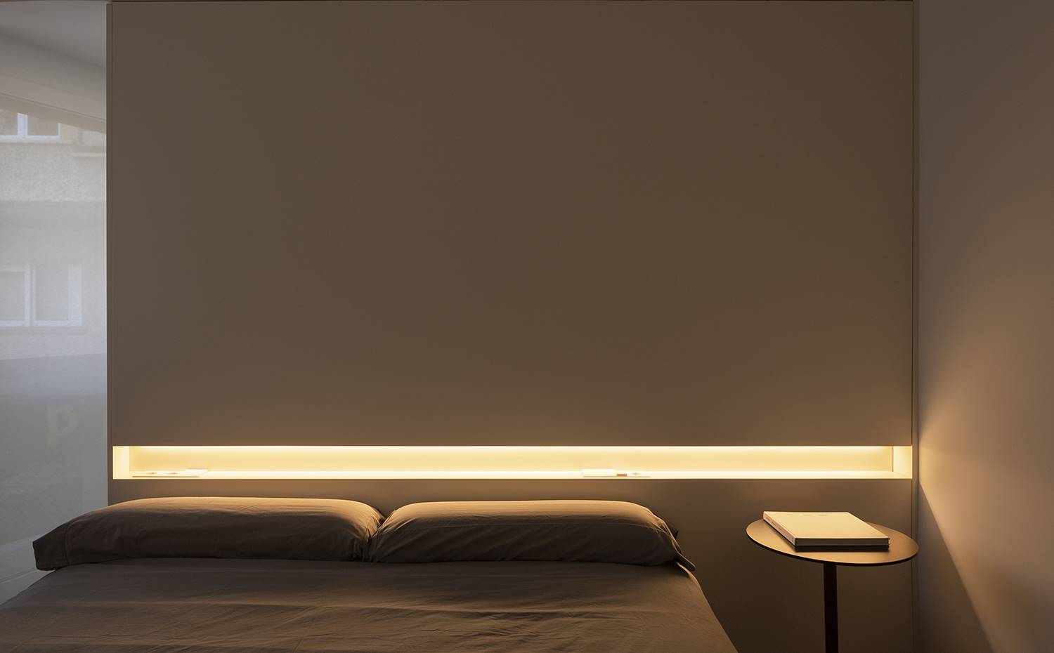 Dormitorio con mesita de noche, libro encima, cabecero con detalle de luz. Dominada por una estética minimalista, el dormitorio principal apuesta por una decoración austera a la vez que efectista y establece un sugerente juego de luces partiendo desde el propio cabecero. 