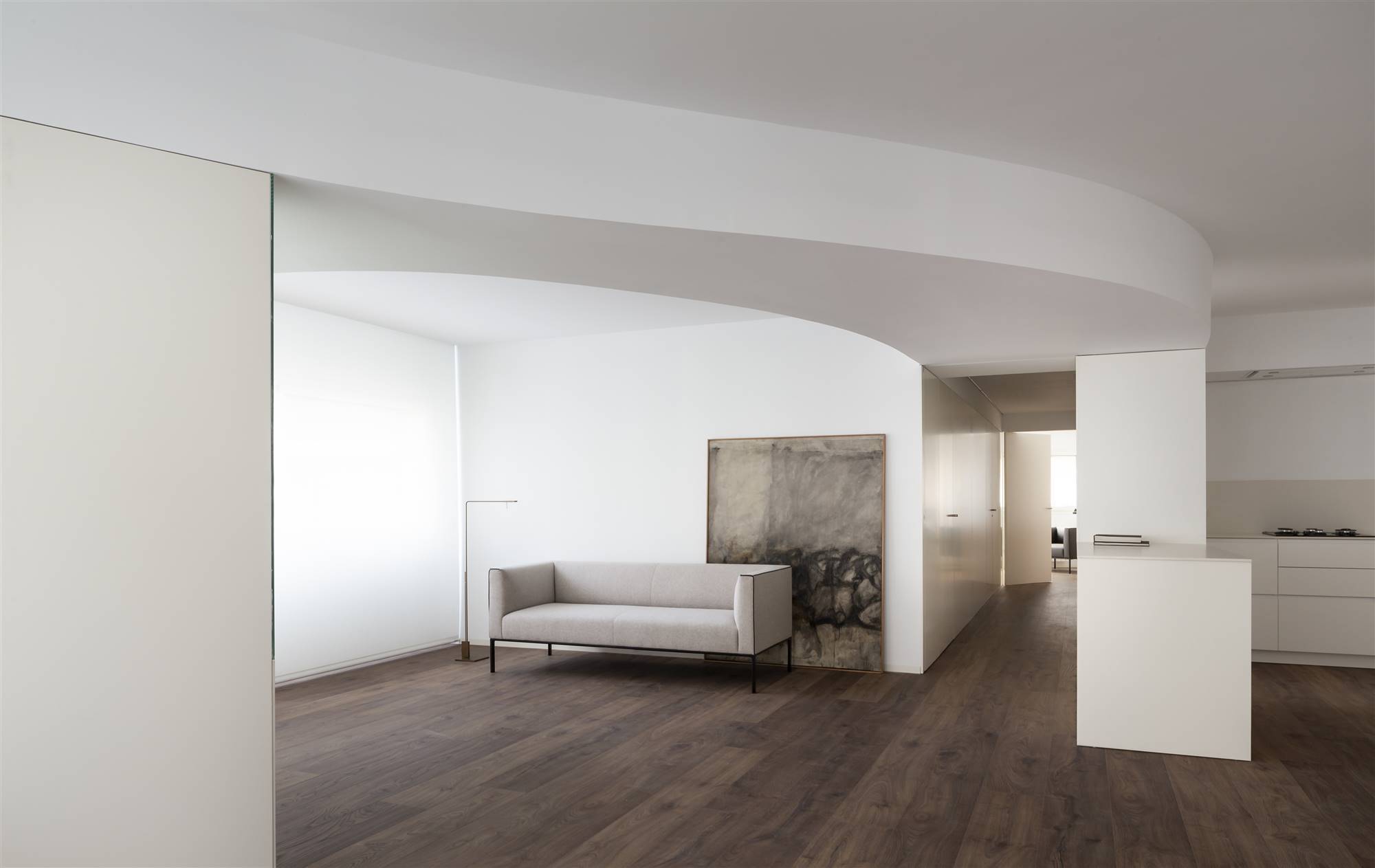 Salón junto a cocina, ambiente diáfano, sofá y cuadro de tonos tierra, techos ondulantes. La curva del techo del salón se crea para separar la zona de cocina y comedor de la de salón, y a su vez para reforzar la idea de diseño integrado, en la que se aunan distintos ambientes en una misma planta diáfana. 