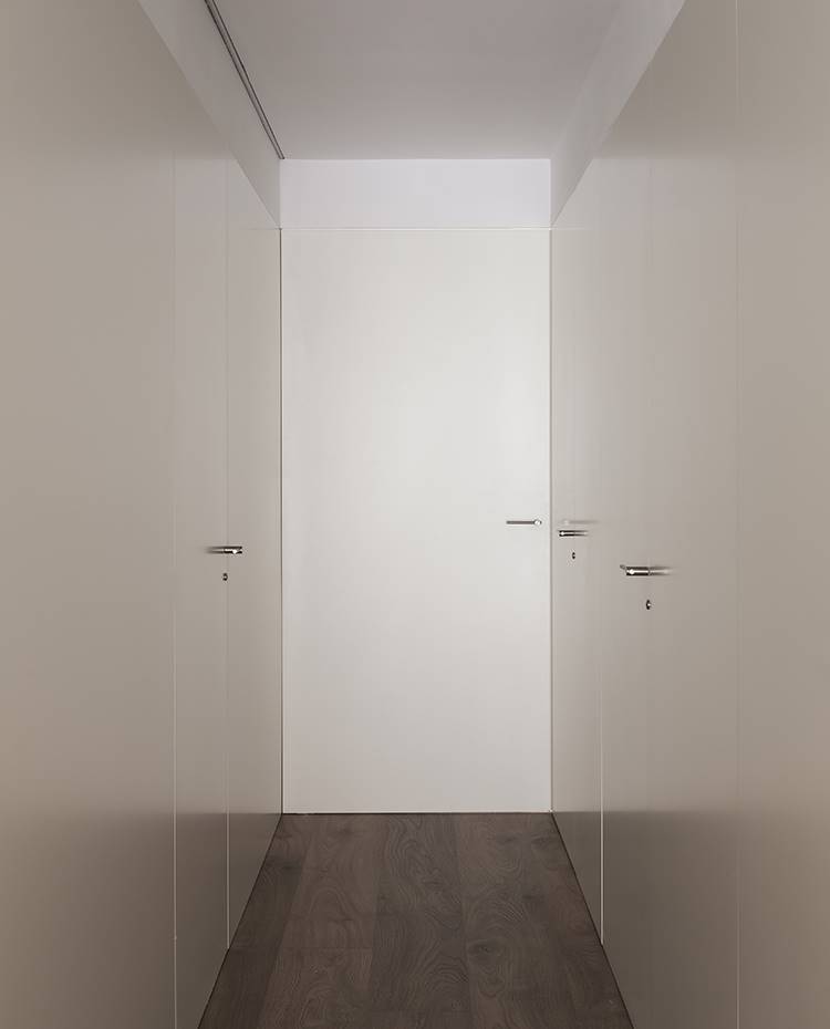 Zona de paso con armarios para el almacenaje en ambos lados. Tras una puerta corredera se accede a un pasillo a través del cual se llega a los dormitorios y baños. A ambos lados se han dispuesto armarios de suelo a techo que brindan una gran superficie de almacenaje sin apenas influir en la estética purista que presenta el proyecto. 