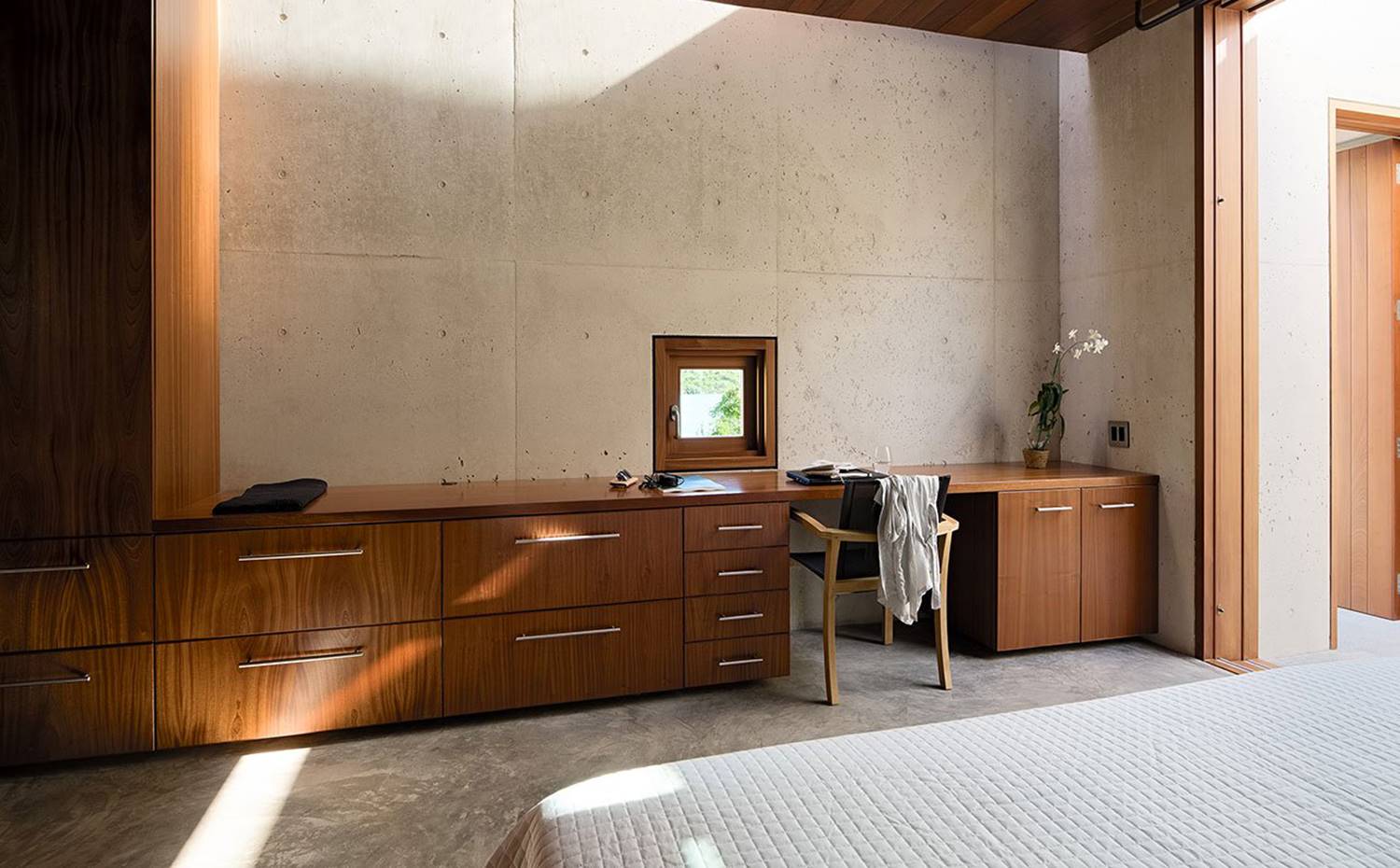 Dormitorio, pared de hormigón, apertura marco en alumino tipo madera, muebles bajos de madera. La decoración interior combina la frescura minimalista de las paredes de hormigón y el pavimento gris con la exótica calidez de la caoba presente en ventanas, puertas y techos.