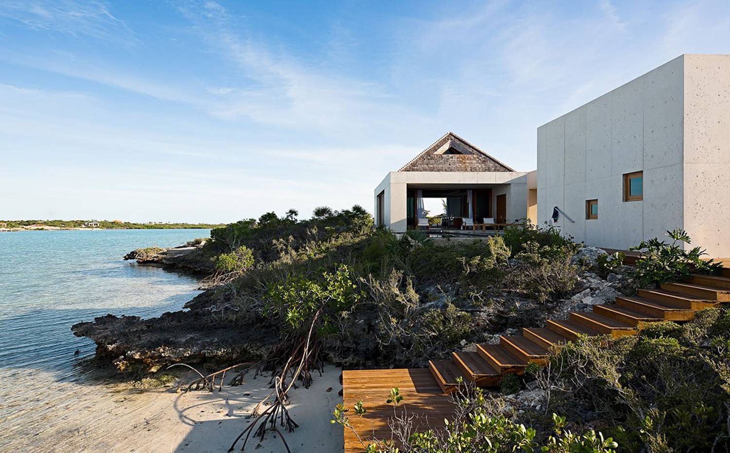 Escaleras de madera voladas que conducen hasta arena de la playa. Estrutura vivienda de hormigón. La vivienda se extiende a lo largo de la costa de las islas Turcas y Caicos con su fachada de hormigón de textura satinada cuyo acabado grisáceo contrasta con el agua del mar turquesa y la flora tropical que cubre el perímetro de la casa.