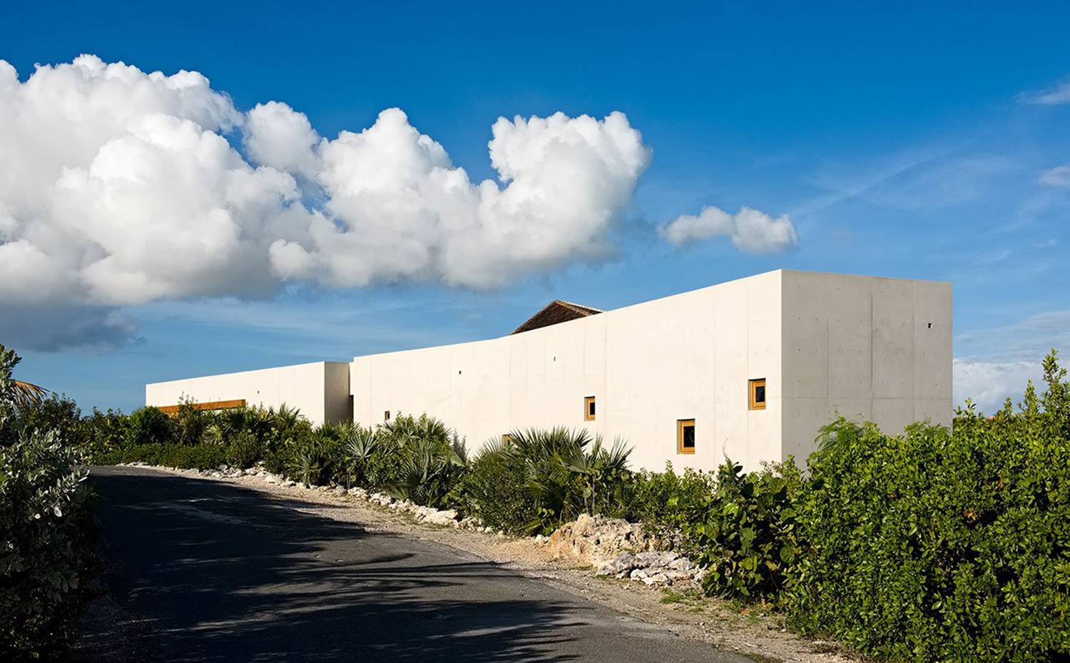 Fachada exterior estructura de hormigón, camino asfaltado, vegetación en arcén. El proyecto llevado a cabo por Rick Joy Architects da la espalda a la calle, formando un baluarte continuo de hormigón, únicamente perforado por unas pocas y pequeñas ventanas cuadradas que dejan entrar la luz preservando la intimidad de sus propietarios.