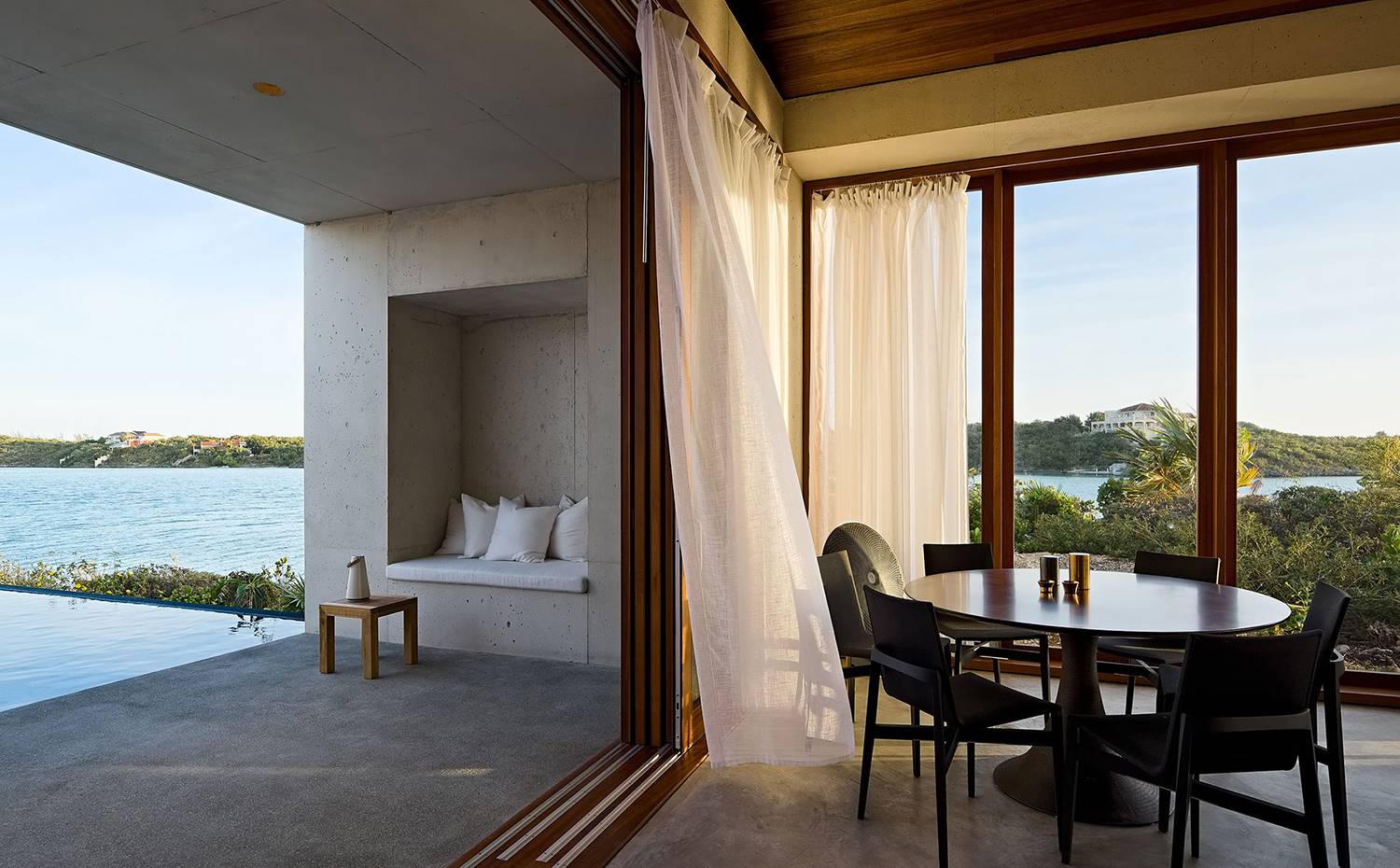 Mesa comedor, cortinas en cerramiento de cristal, mesita auxilar madera, blanco de obra integrado en estructura de hormigón. Desde el interior, los espacios se sienten aislados y protegidos, y las vistas de la bahía desde el pabellón donde se ubica el salon-comedor y la cocina parecen totalmente exclusivas. 