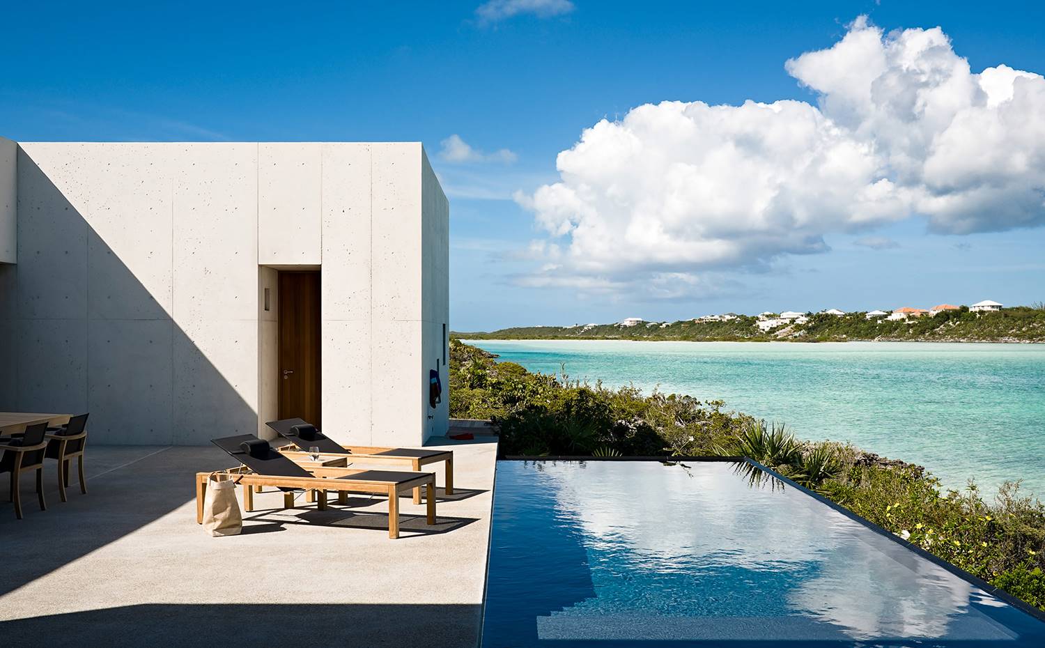 Tumbonas de madera junto a la piscina infinita asoman hacia el mar. En la parte exterior una piscina infinita poco profunda crea una transición entre la arena de la playa y la terraza adyacente, acercando la extensión del agua del océano a los espacios habitables.