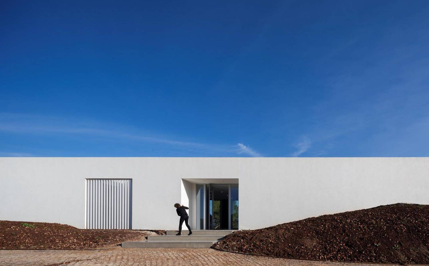 Entrada vivienda, montículo tierra entorno vivienda, escaleras de acceso, camino empedrado. En la fachada oeste se ubica la puerta de acceso a la vivienda, convirtiéndose en la única apertura en este lado de la casa y preservando así la privacidad ante el camino de acceso a la casa. 