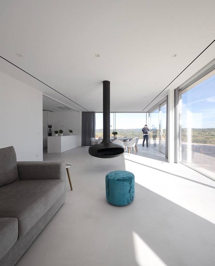 Salón con sofá gris, puf aterciopelado turquesa, chimenea de acero negra, mesas blancas comedor. En el extremo de la vivienda se emplaza el salón, junto a comedor y cocina, conectado visual y físicamente al exterior a través de cerramientos acristalados que van de techo a suelo, y que a su vez se abren hacia la terraza continua.
