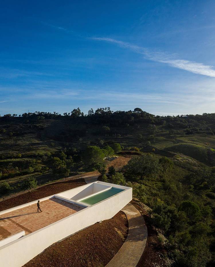 Vista de la cubierta apoyada sobre montículode tierra, paisaje entorno vegetación  . Las formas rectas y puntas afiladas de la estructura de la vivienda se enfatizan con el blanco inmaculado de la fachada y que da forma a un cuerpo de estética depurada que contrasta con los tonos tierra de su base y el intenso verdor de su entorno.
