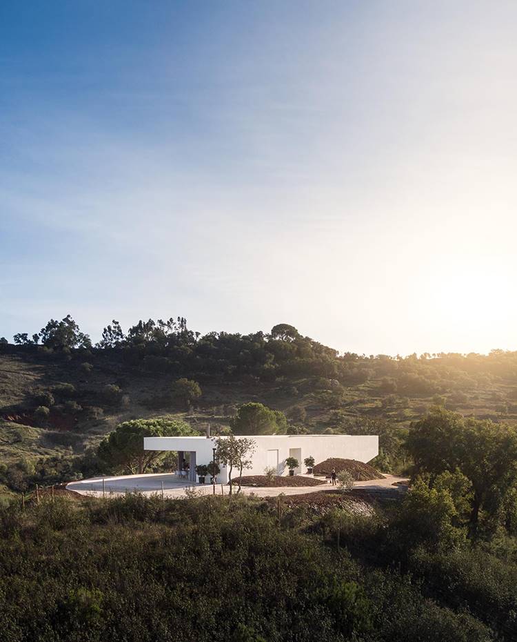 Vista vivienda entre entorno natural, estructura cúbica en blanco. La vivienda, situada en las colinas del sur de Portugal, concretamente en el municipio de São Bartolomeu de Messines, se erige como un moderno monumento en el paisaje rural, creación del estudio portugués Vitor Vilhena Arquitectura.