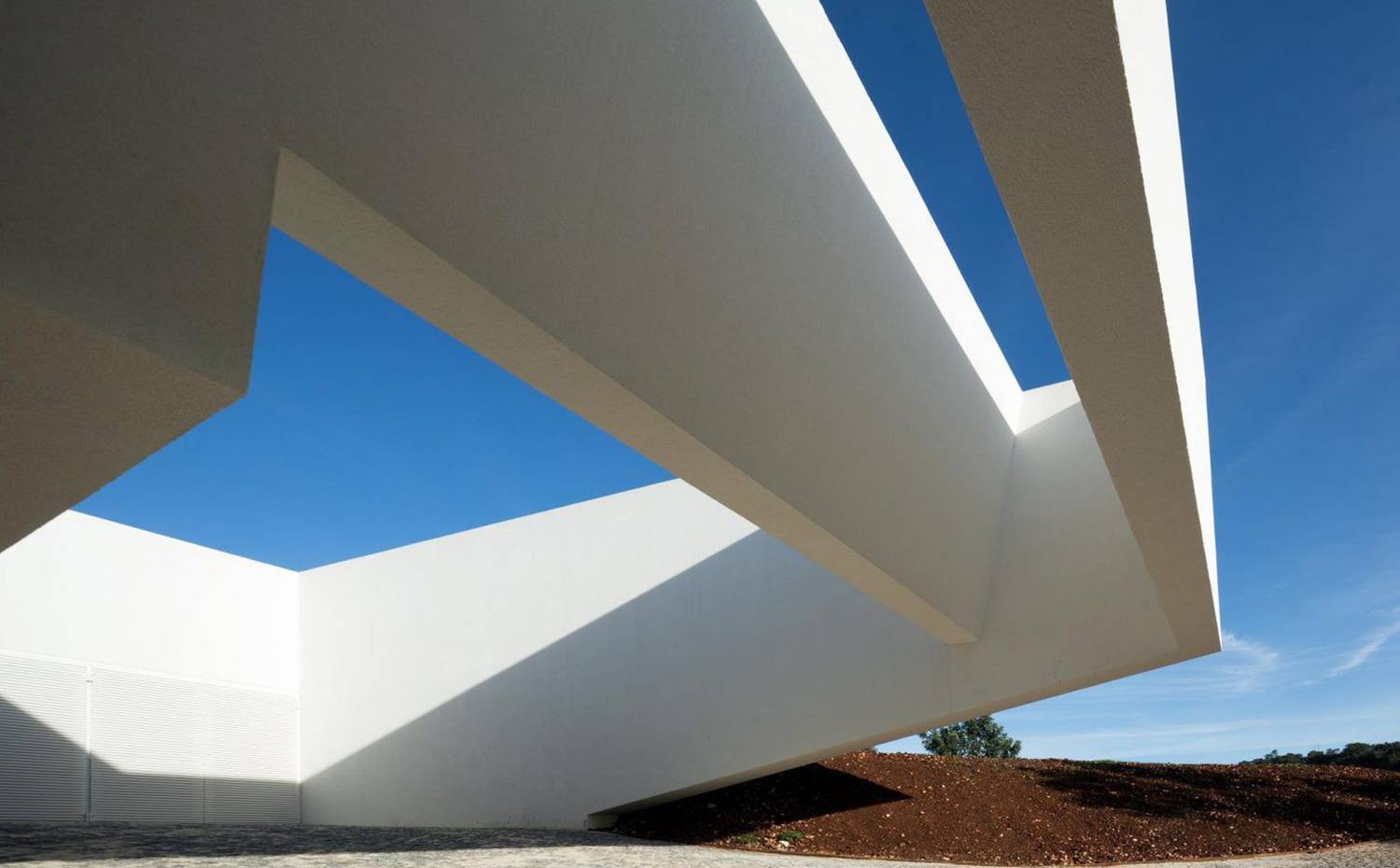 Voladizo del edificio sobre el suelo con aperturas cenitales . Toda la escultórica estructura exterior de la vivienda contrasta con el encantador entorno de la campiña portuguesa. Su imponente fachada blanca mate descansa en una privilegiada ubicación.  