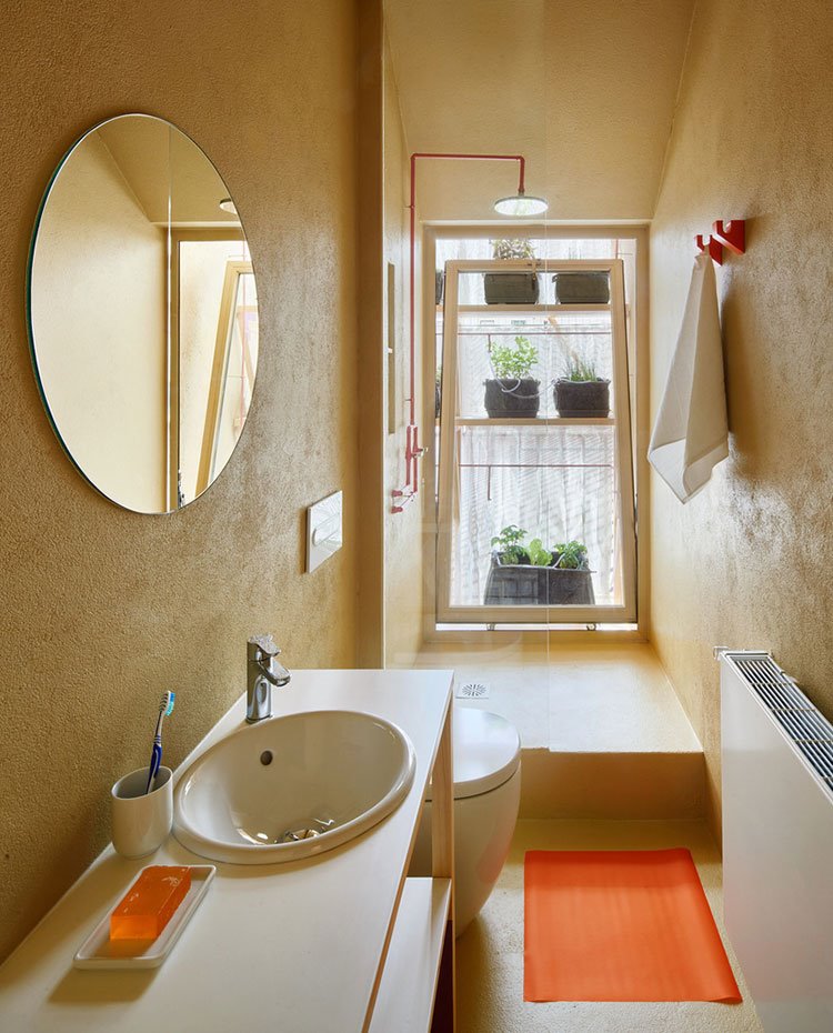 Baño con lavamanos encastrado en encimera sobre estructura abierta, espejo circular, alfombra naranja, plato de ducha de obra con hoja de cristal transparente, apertura hacia jardín 
