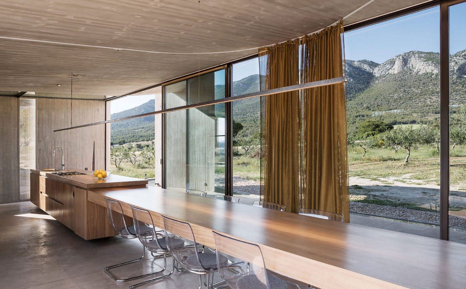 Cocina con comedor, isla de madera, mesa de centro continua de madera y gran capacidad, sillas transparente, vista exterior a campo y montañas, cortinas mostaza.