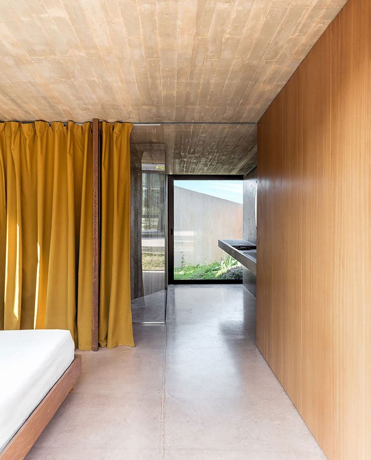 Dormitorio con estructura de madera, revestimiento pared madera, suelo de hormigón, cerramiento cristal transparente a bajo, encimera de piedra suspendida, cortinas mostaza