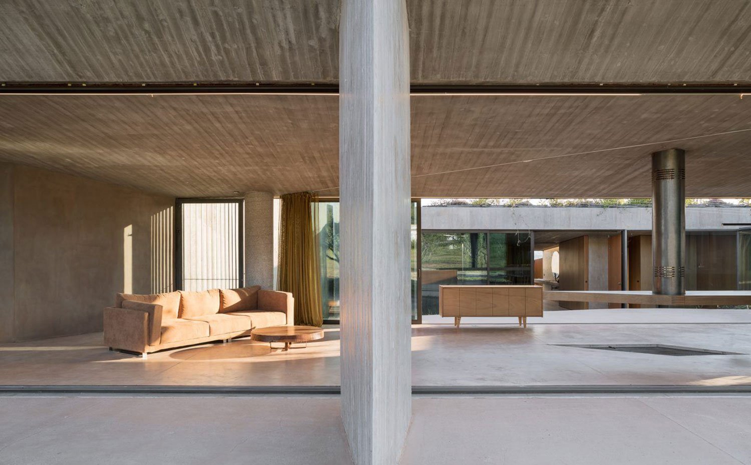 Estructura exterior vivienda, con cerramientos abiertos, pavimento continuo de hormigón, sofá marrón, mesita circular madera, consola madera, chimenea suspendida.