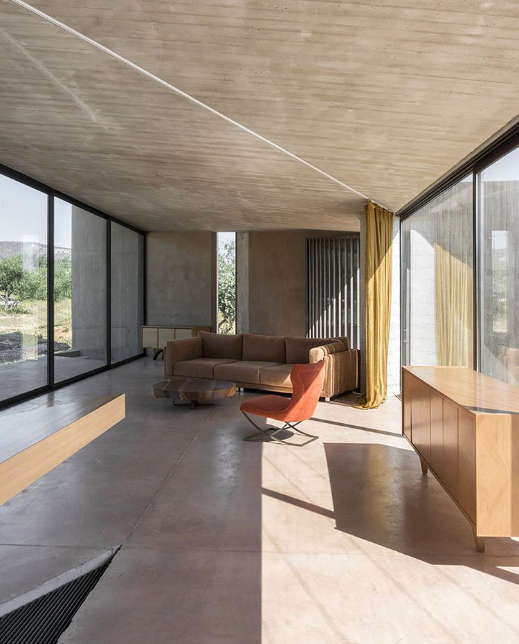 Salón con suelo de hormigón, módulo consola de madera, butaca naranja, mesa de centro circular, sofá marrón, cortinas de gran altura de terciopelo mostaza, cerramientos acristalados