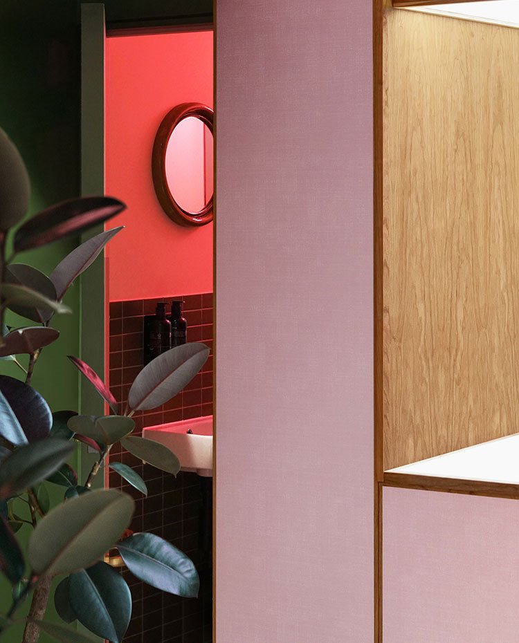 Acceso baño con módulo de madera rosa y natural, pared pintada de verde, planta y pared baño revestida de baldosas negras en parte baja con iluminación en tonos rosas