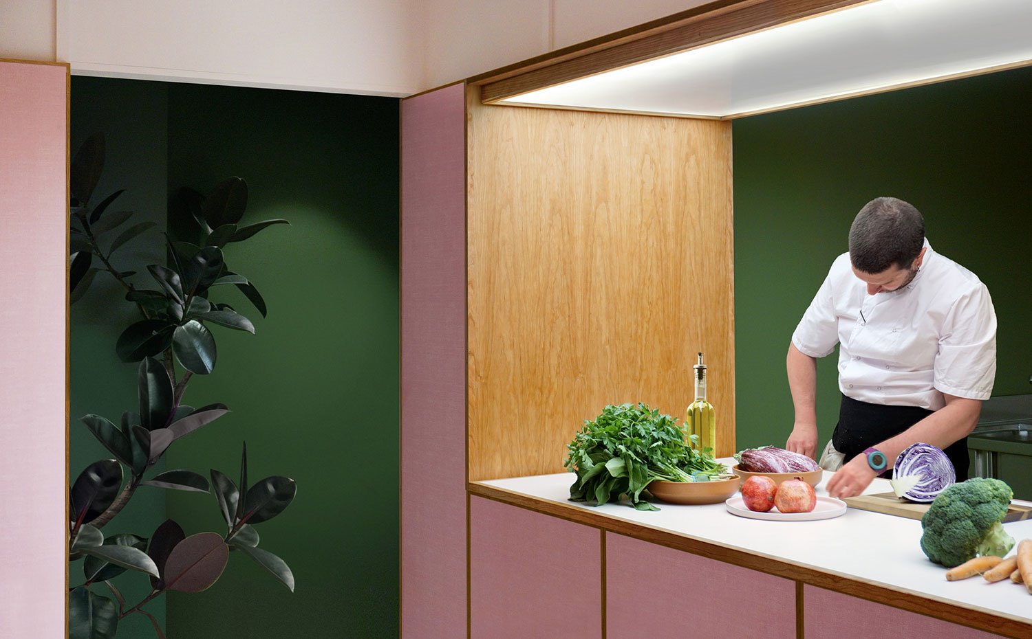 Mostrador cocina abierta con sobre en madera blanca, estructura en combinación madera natural y frentes en rosa, cocinero con vegetales, plafón superior bajotecho, pared de fondo en verde