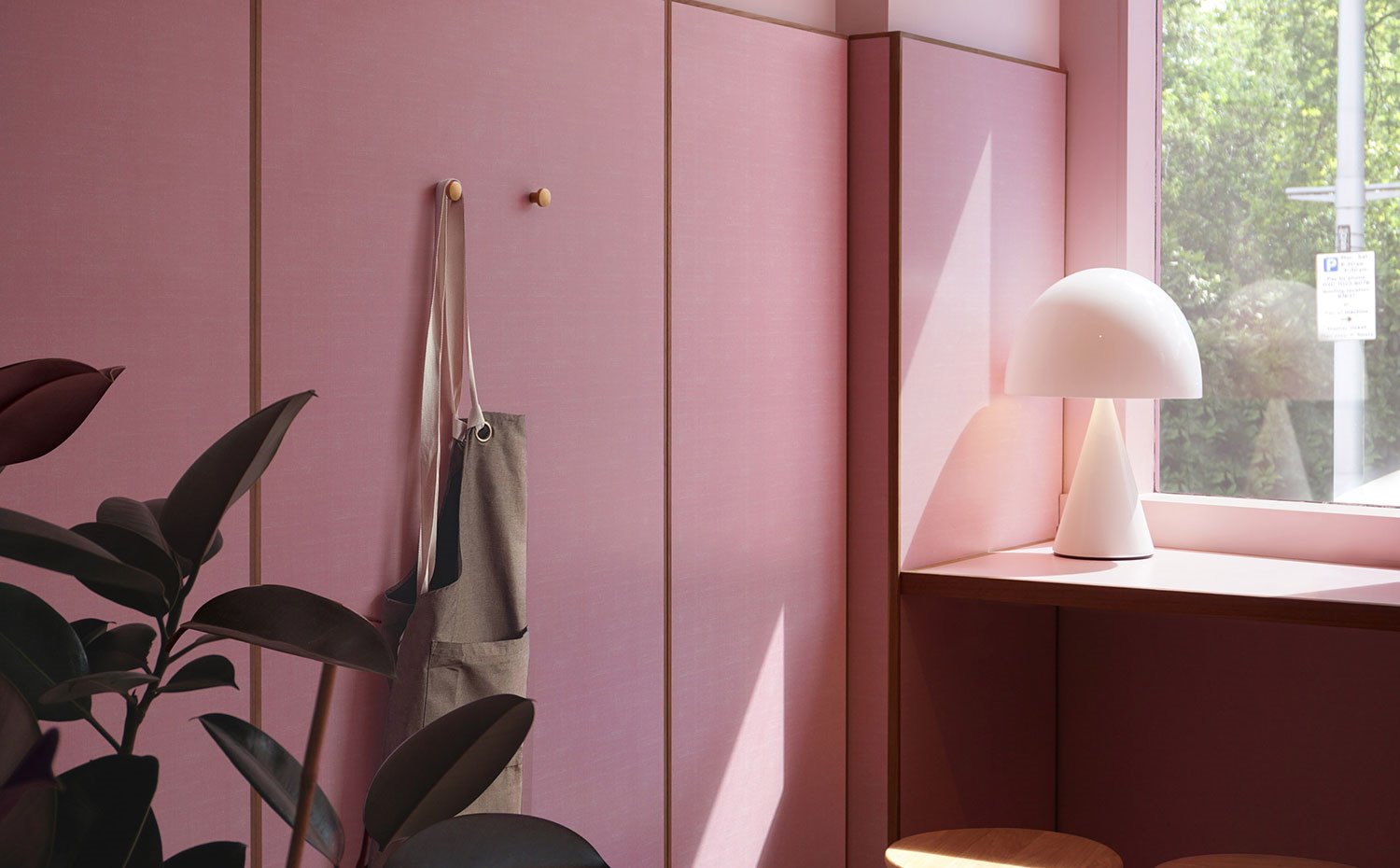 Zona ventana a calle con barra, luminaria de sobremesa blanca, taburete de madera, panel de madera en rosa con marco natural, colgador dorado, delantal marrón