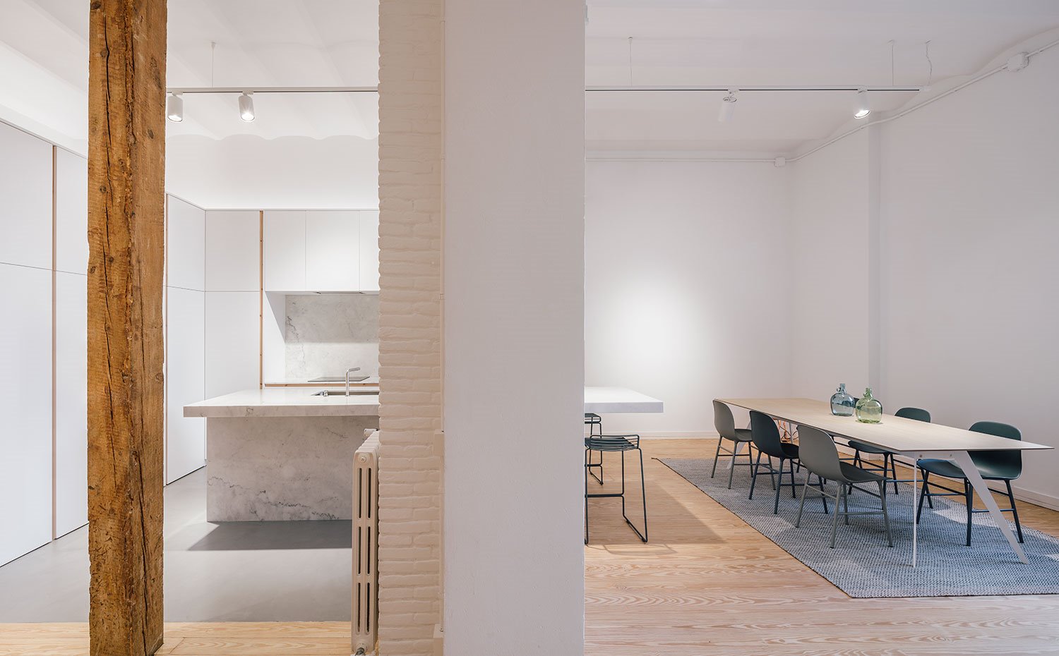 Cocina-con-isla-y-muebles-blancos-continuos,-radiador-en-pared-de-obra,-pilar-madera,-mesa-comedor-de-acero-y-sobre-de-madera,-sillas-grises-y-alfombra-de-fibra-paredes-en-blanco